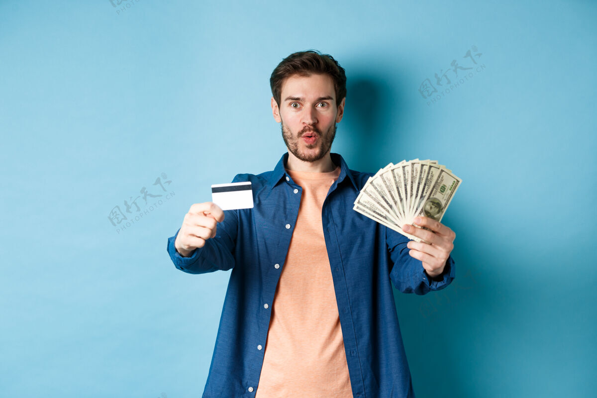 人兴奋的现代人展示着钞票和塑料信用卡 站在蓝色的背景上说“哇哦 太棒了”成人财务男性