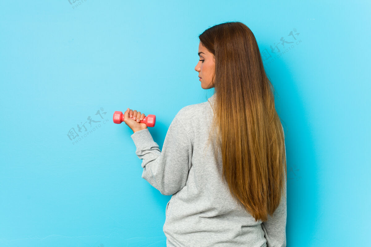 运动一个年轻的白人妇女拿着一个被隔离在蓝色表面上的哑铃活力健身女性