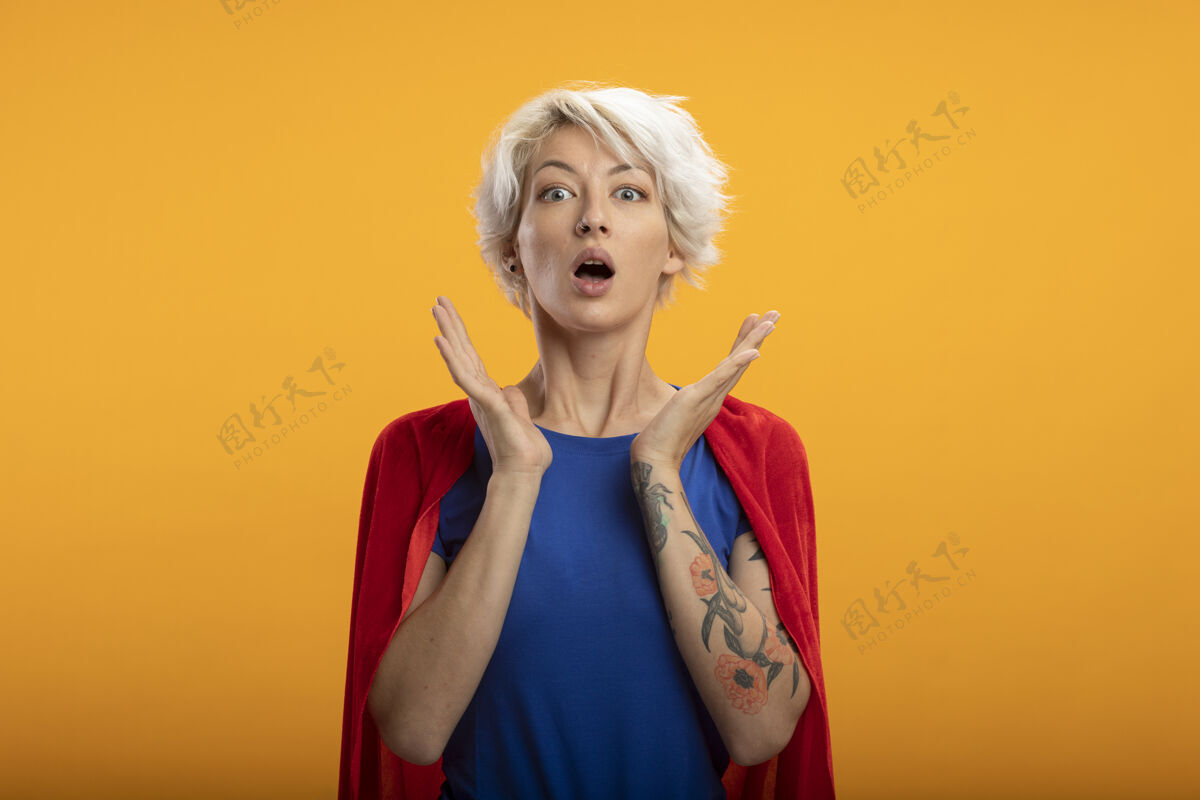 表情震惊的女超人穿着红色斗篷 举起双手孤立地站在橙色的墙上人姿势超级英雄