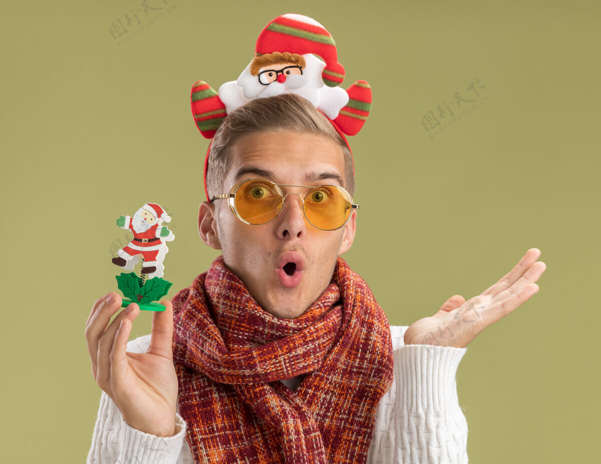 显示令人印象深刻的年轻帅哥戴着圣诞老人头带和围巾看着相机拿着圣诞老人玩具显示空手孤立在橄榄绿的背景圣诞老人家伙绿色