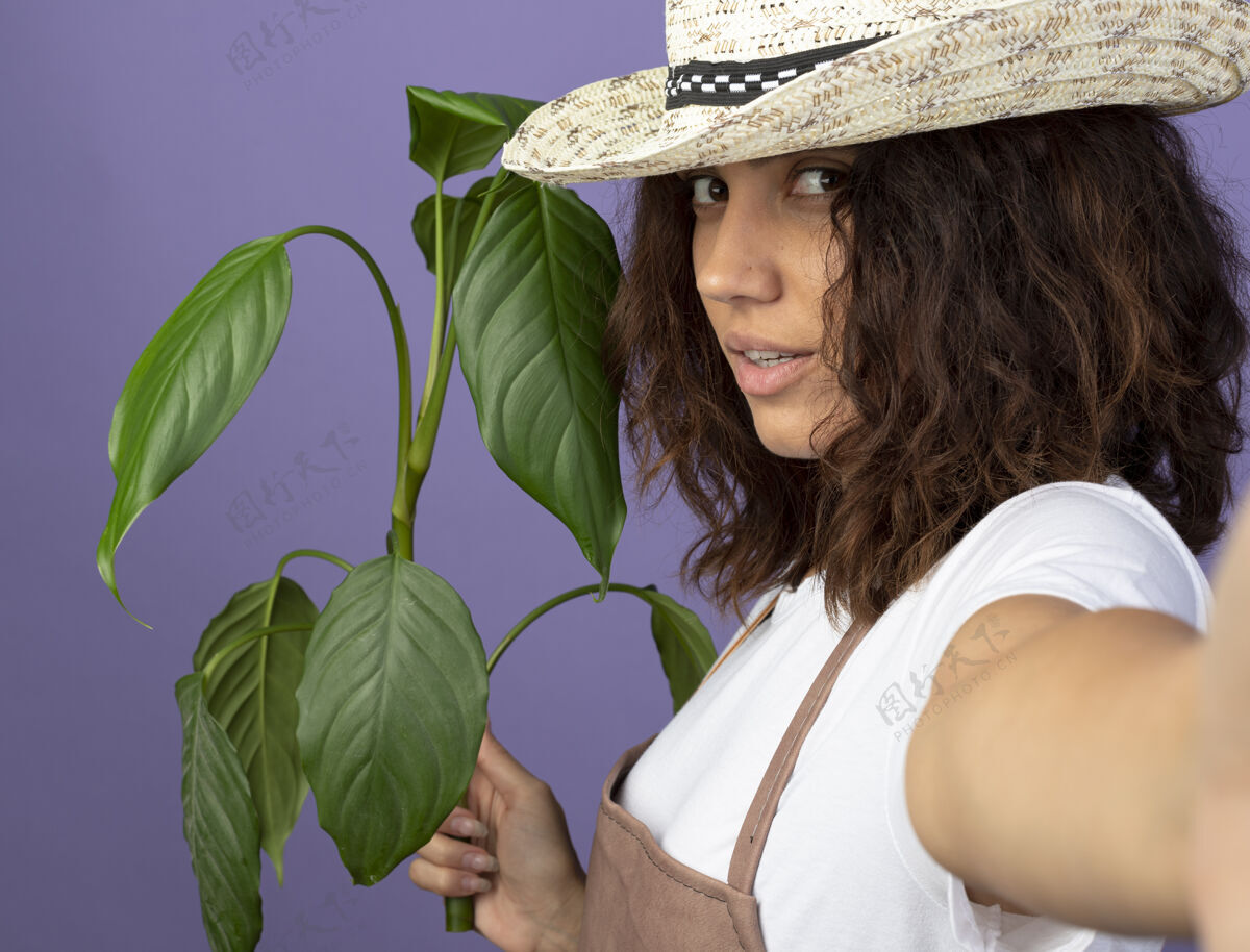 信心自信的年轻女园丁穿着制服 戴着园艺帽 拿着一株紫色隔离带上的相机举行花园紫色