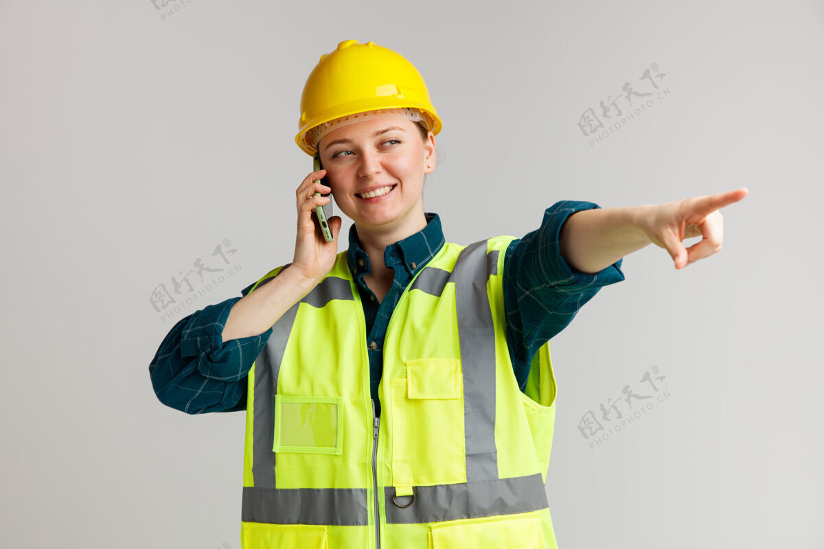 微笑面带微笑的年轻女建筑工人戴着安全帽和安全背心打电话看着并指着旁边头盔电话建筑