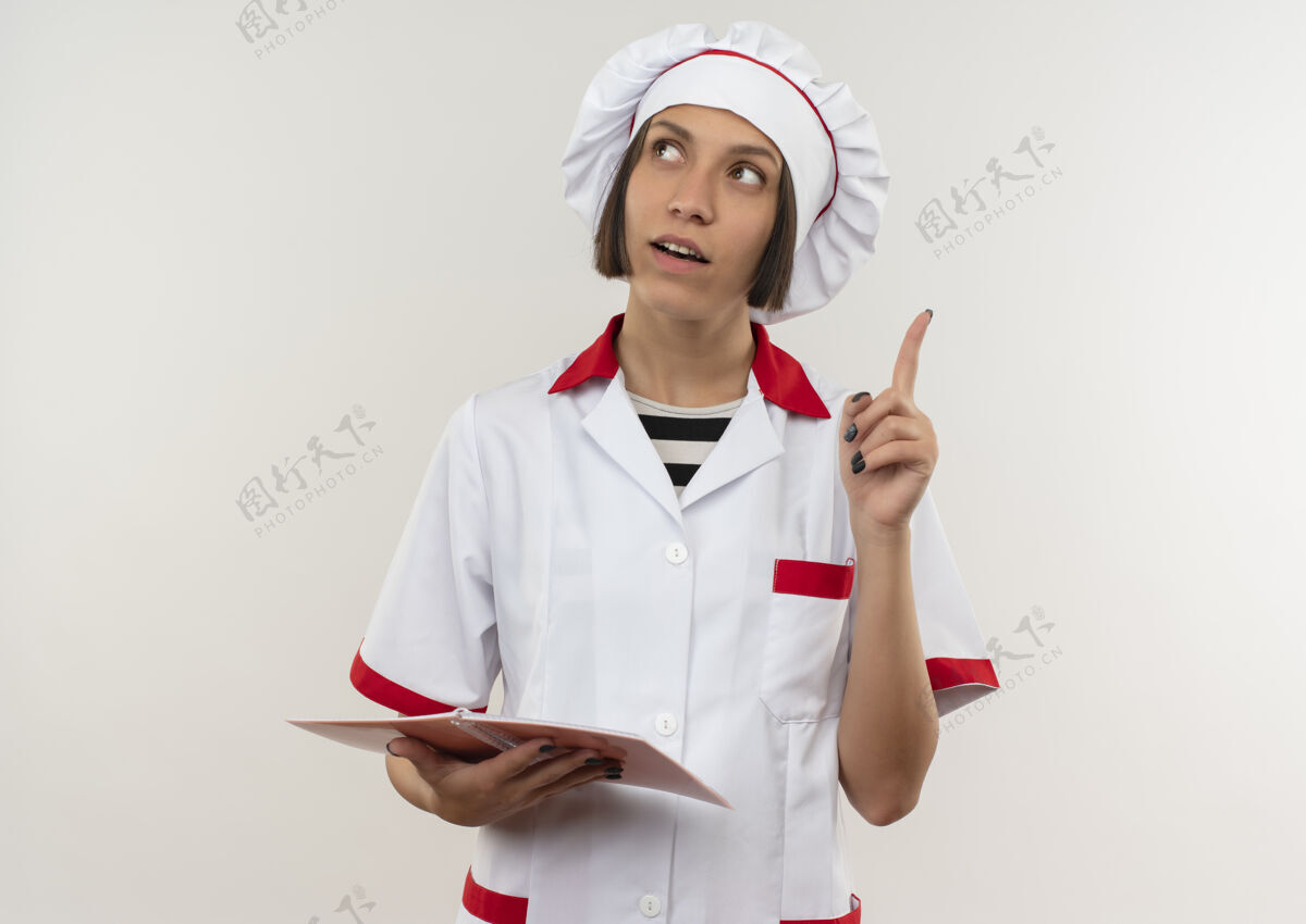 空间印象深刻的年轻女厨师在厨师制服举行记事本和提高手指看一边孤立的白色与复制空间厨师制服笔记