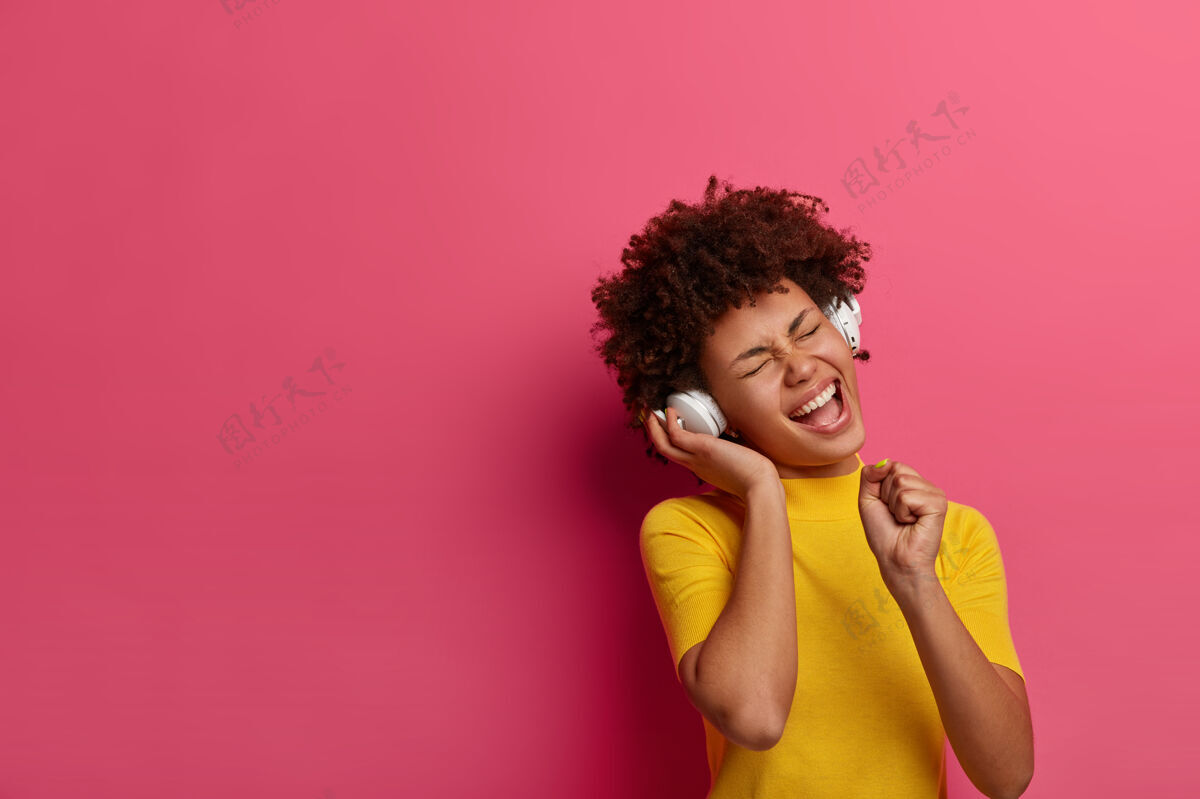 青少年是的 很棒的声音欣喜若狂的卷发女人用耳机听音频 握紧拳头拿着麦克风 闭上眼睛 穿着黄色t恤 带着良好的寒意 孤立在粉红色的墙上人艺术家耳机