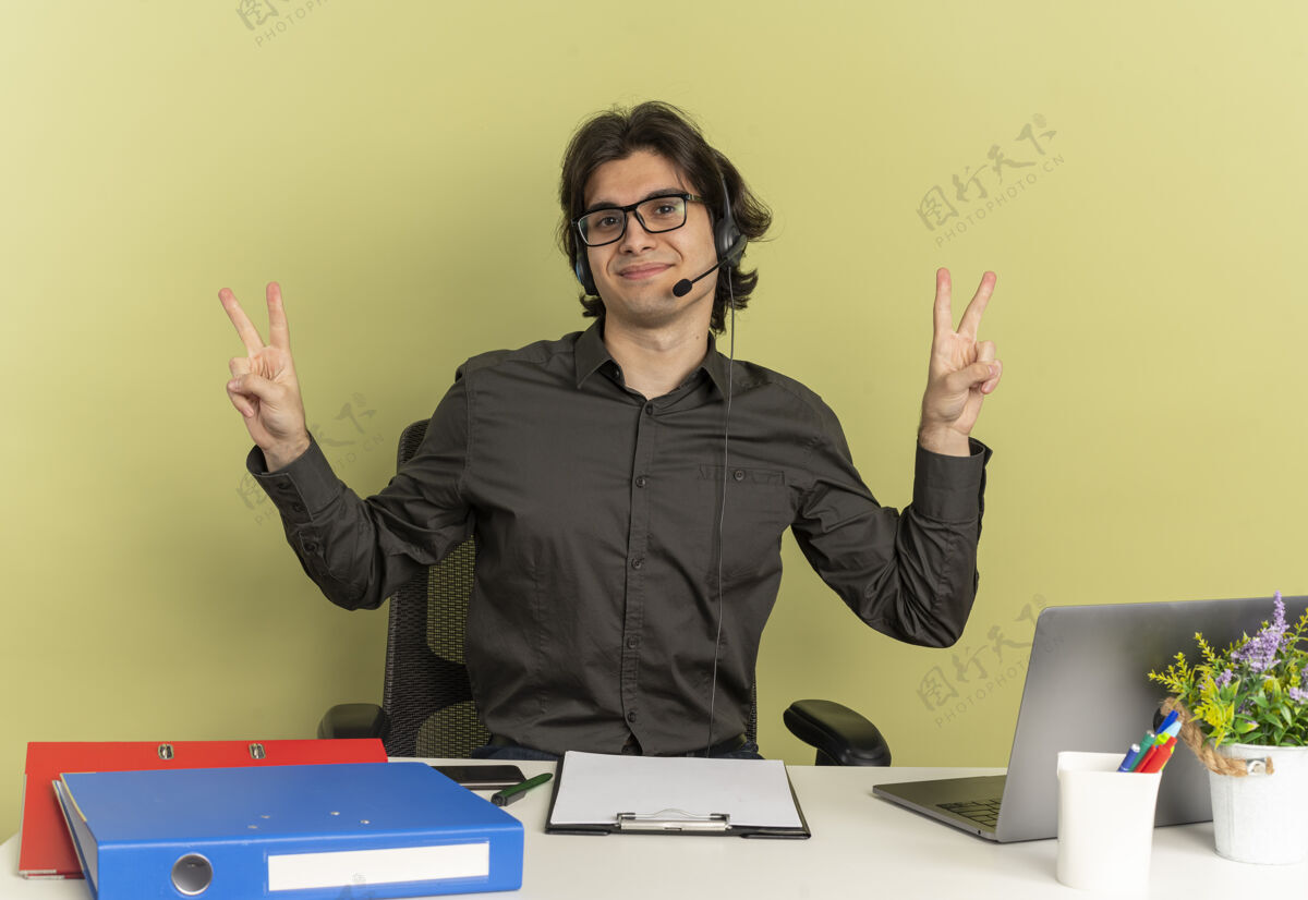 手戴着耳机 戴着眼镜的年轻上班族坐在办公桌旁 拿着办公工具 用笔记本电脑和手势 胜利手势被隔离在绿色背景上 还有复印空间眼镜笔记本电脑办公室
