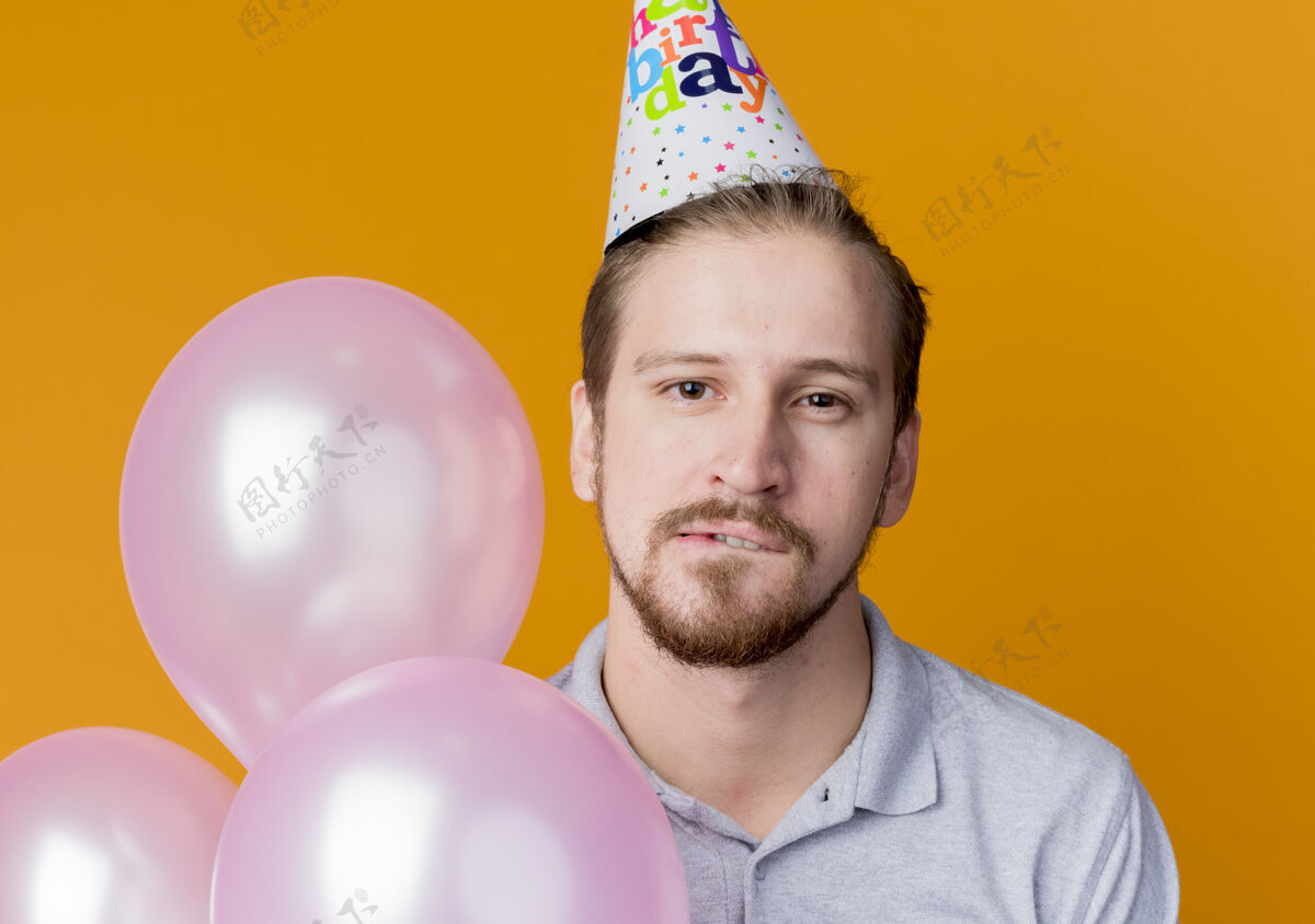 咬戴着派对帽的迷人年轻人手持一束气球咬着嘴唇站在橙色的墙上生日派对的概念站嘴唇看