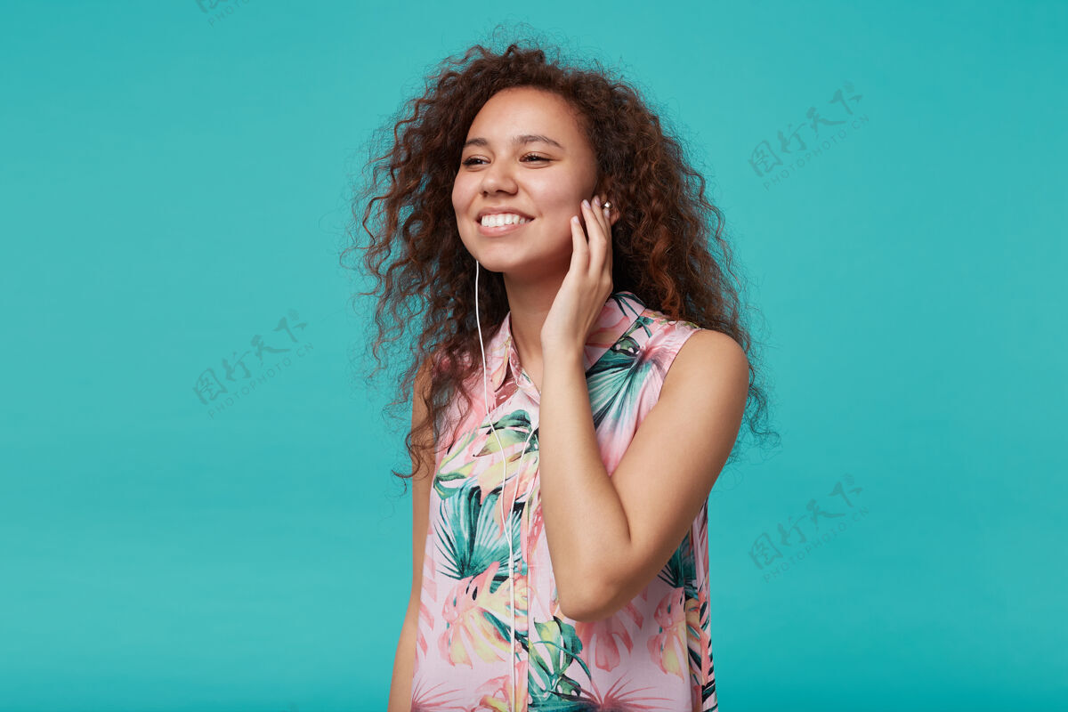 女士积极向上的年轻可爱的棕色卷发女士 随意的发型 举起手放在听筒上 微笑着欣赏喜爱的音乐曲目 与蓝色隔绝女人花漂亮