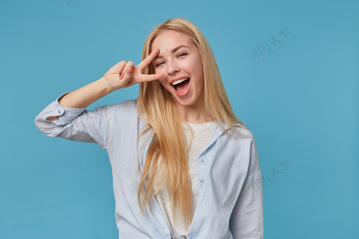 宽快乐的年轻金发女性的肖像 长发 穿着蓝色衬衫和灰色t恤 站着的时候用和平的手势握着手指 面带微笑头发女性表情