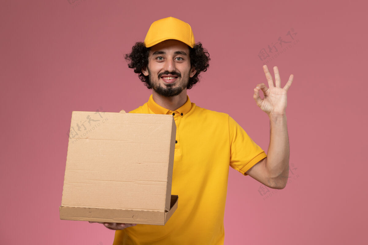 男正面图：身穿黄色制服的男快递员拿着食品快递箱站在浅粉色的墙上工作持有盒子