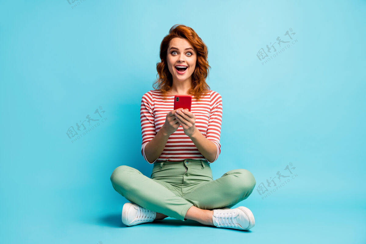 快乐全尺寸照片的热情惊讶的女孩坐在双腿交叉使用手机阅读社会网络新闻印象深刻尖叫哇omg穿绿白色鞋子蓝色隔离年轻地板红发