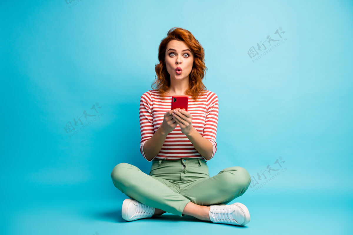 情绪全身照片惊讶的女孩坐在双腿交叉使用手机阅读社会媒体信息印象深刻尖叫哇omg穿时尚的衣服蓝色隔离手机短信腿