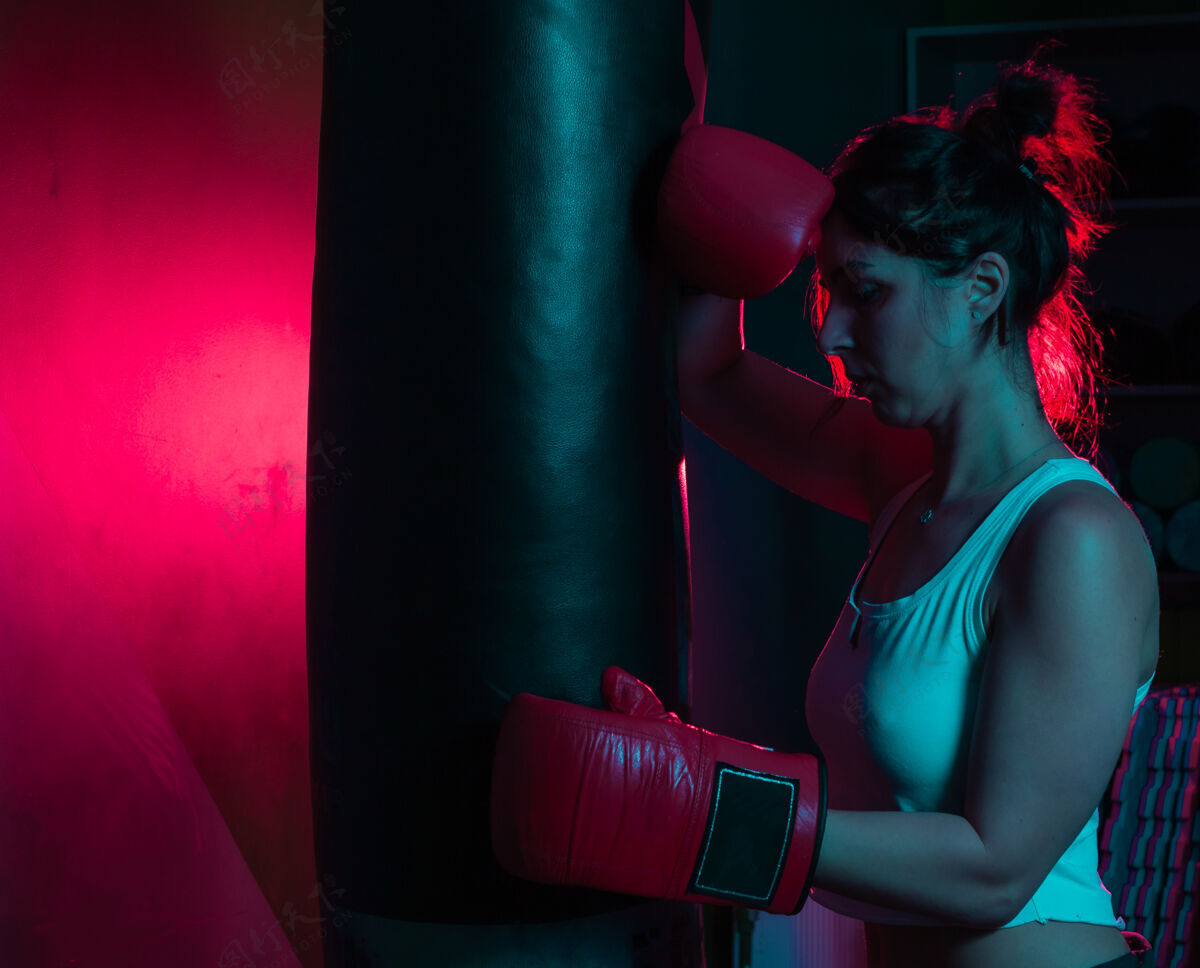 拳击手戴着拳击手套的疲惫的女拳击手正靠在一个拳击袋上 背景是红蓝色的霓虹灯肌肉女子盒子