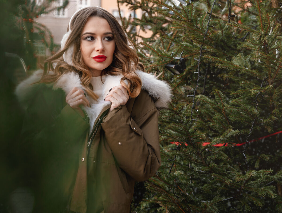 情感穿着针织毛衣和蓬松耳机的白人女士户外写真年轻美丽快乐的微笑女孩穿着皮草外套在装饰圣诞树附近波兰黑发有趣青少年