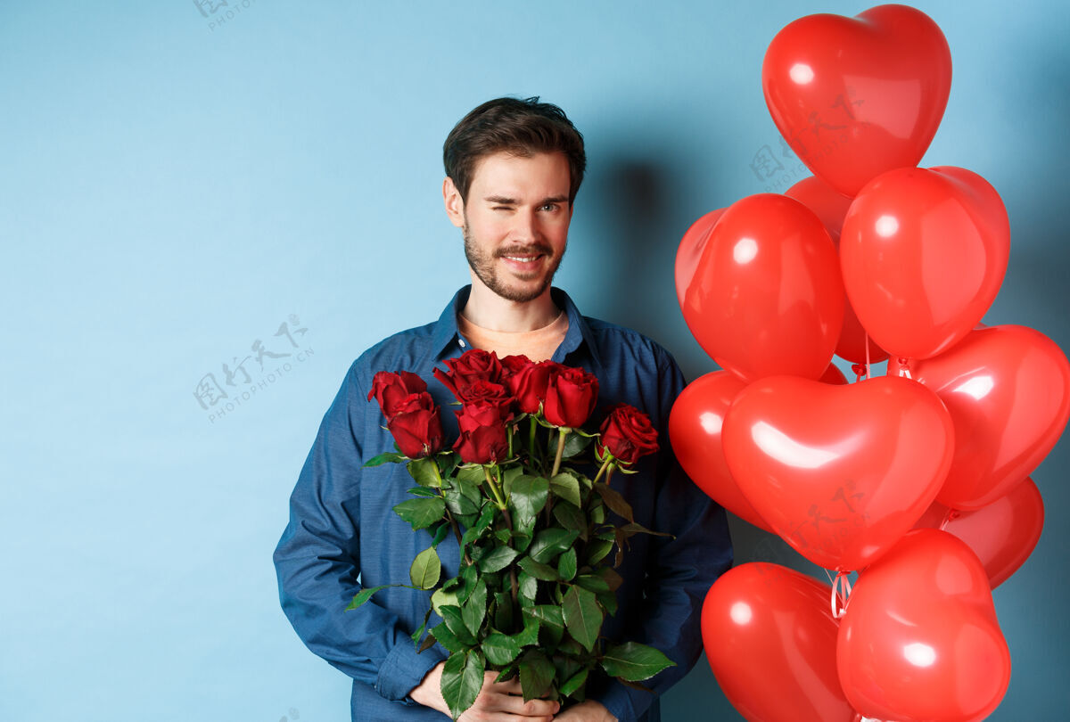 微笑浪漫的男友眨眼微笑 在情人节捧着花束 站在情人的心气球旁 蓝色背景情感年轻花