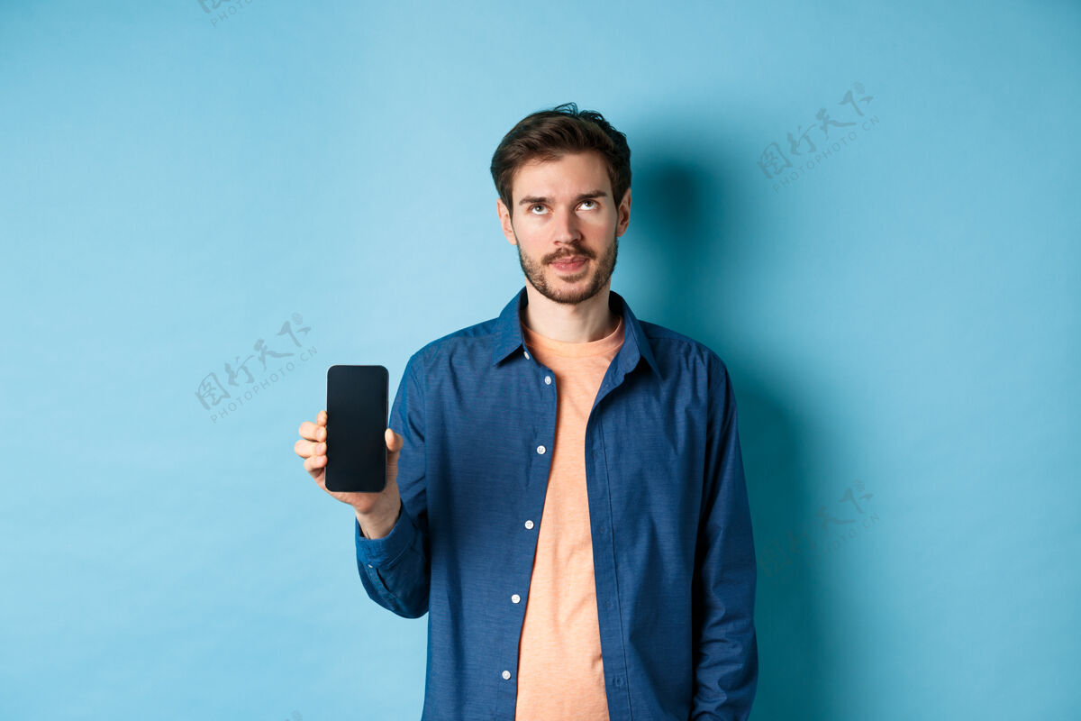 手恼怒的年轻人显示空智能手机屏幕和卷起眼睛困扰 站在蓝色的背景移动朋友交谈