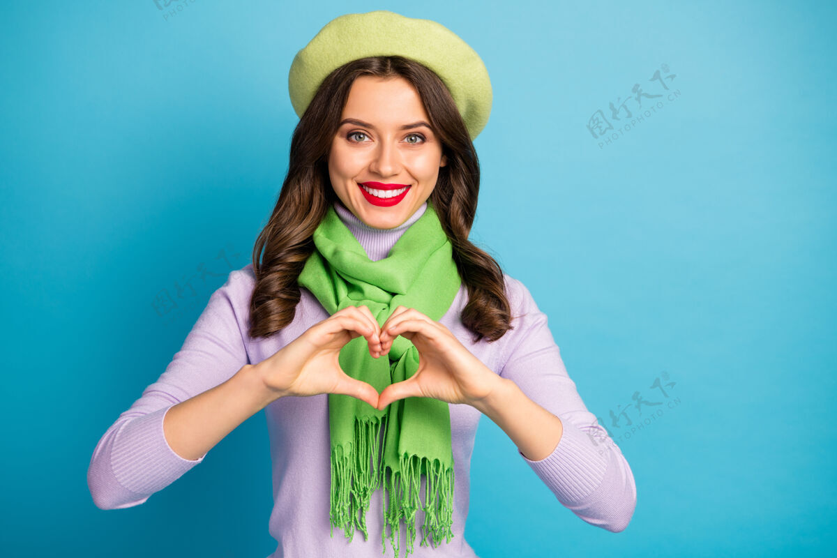 时尚照片中美丽迷人的女士手牵手心形象征心意真挚的感情穿上时尚的绿色贝雷帽紫色高领围巾隔离蓝色的墙壁帽子情感情人