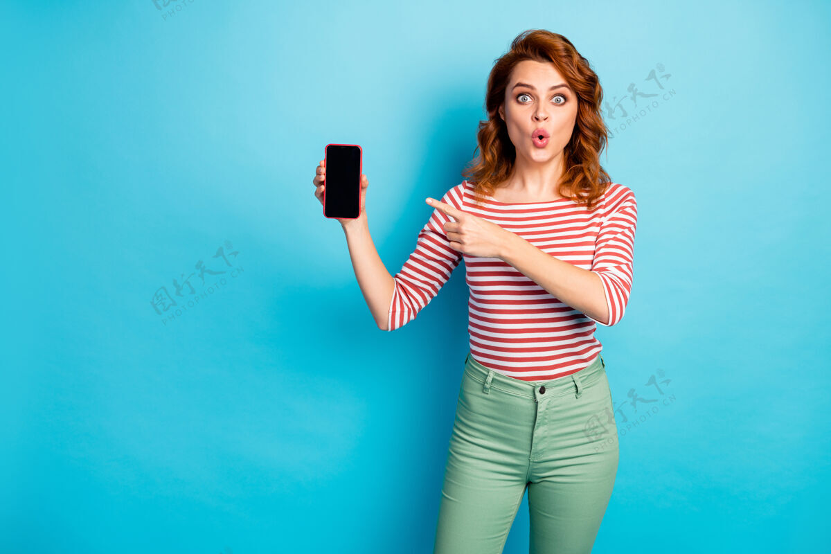 惊喜看新的现代设备惊呆了疯狂的女人拿着智能手机点食指表示难以置信的广告促销印象深刻尖叫哇omg穿套头衫孤立的蓝色生活方式时尚套头衫