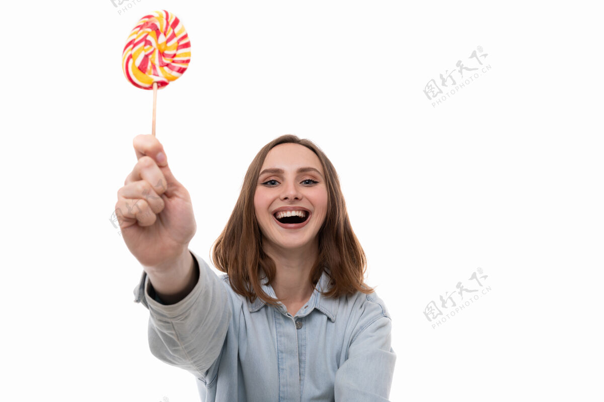 美丽一位身穿牛仔衬衫 面带微笑的年轻女子在白色背景上摆出一个彩色棒棒糖的姿势魅力肖像焦糖