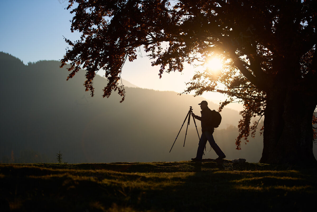 探索徒步旅行者带着相机在大树下的山景背景下的草谷里范围夜晚爱好