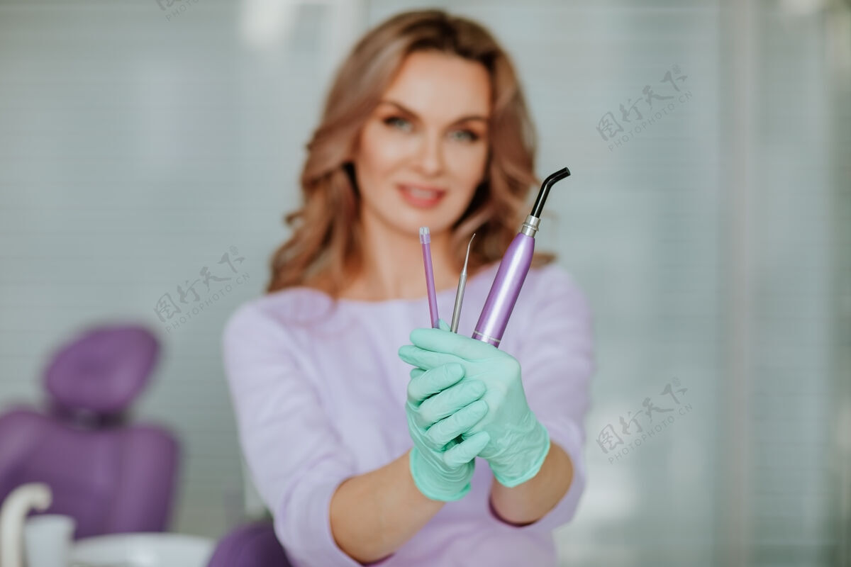 专家年轻迷人的牙医肖像 长卷发 身着紫色医疗制服 戴着绿色医疗手套 在柜子里摆着工具的姿势肖像美白制服