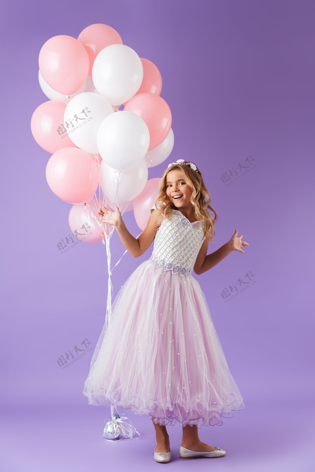 小一个穿着公主裙的漂亮小女孩孤零零地站在紫罗兰色的墙上 手里拿着一堆气球束庆祝连衣裙