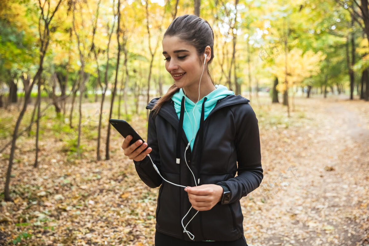 电话微笑的健身女 戴着耳机听音乐 手持手机站在公园里成人跑步女运动员