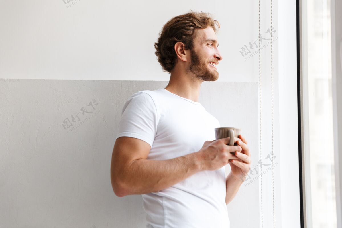 早餐微笑的年轻人端着一杯茶 站在家里的窗前年轻咖啡室内