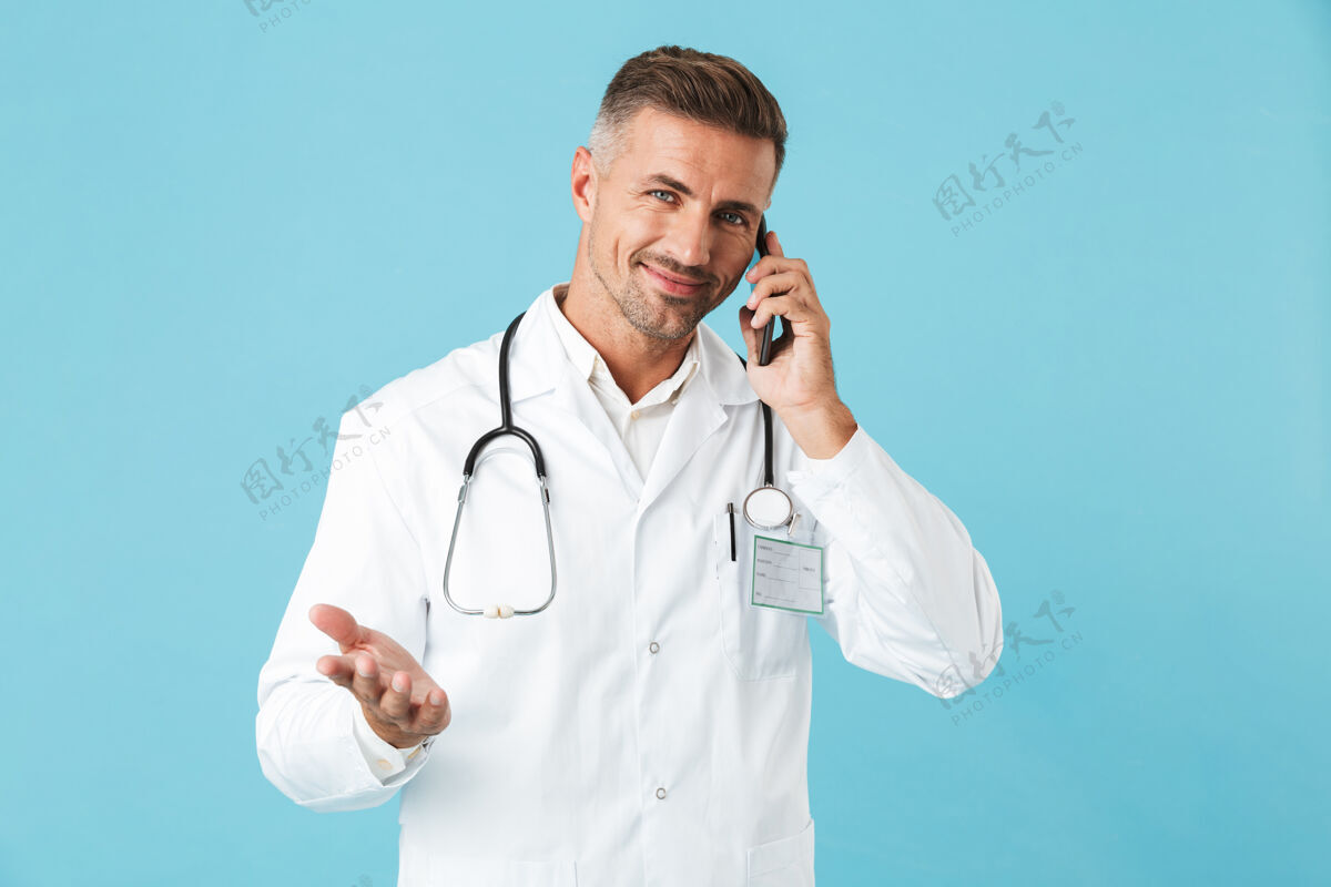 男人快乐的医生穿着白大褂 手持听诊器讲着手机 孤零零地站在蓝色的墙上谈话治疗中年人