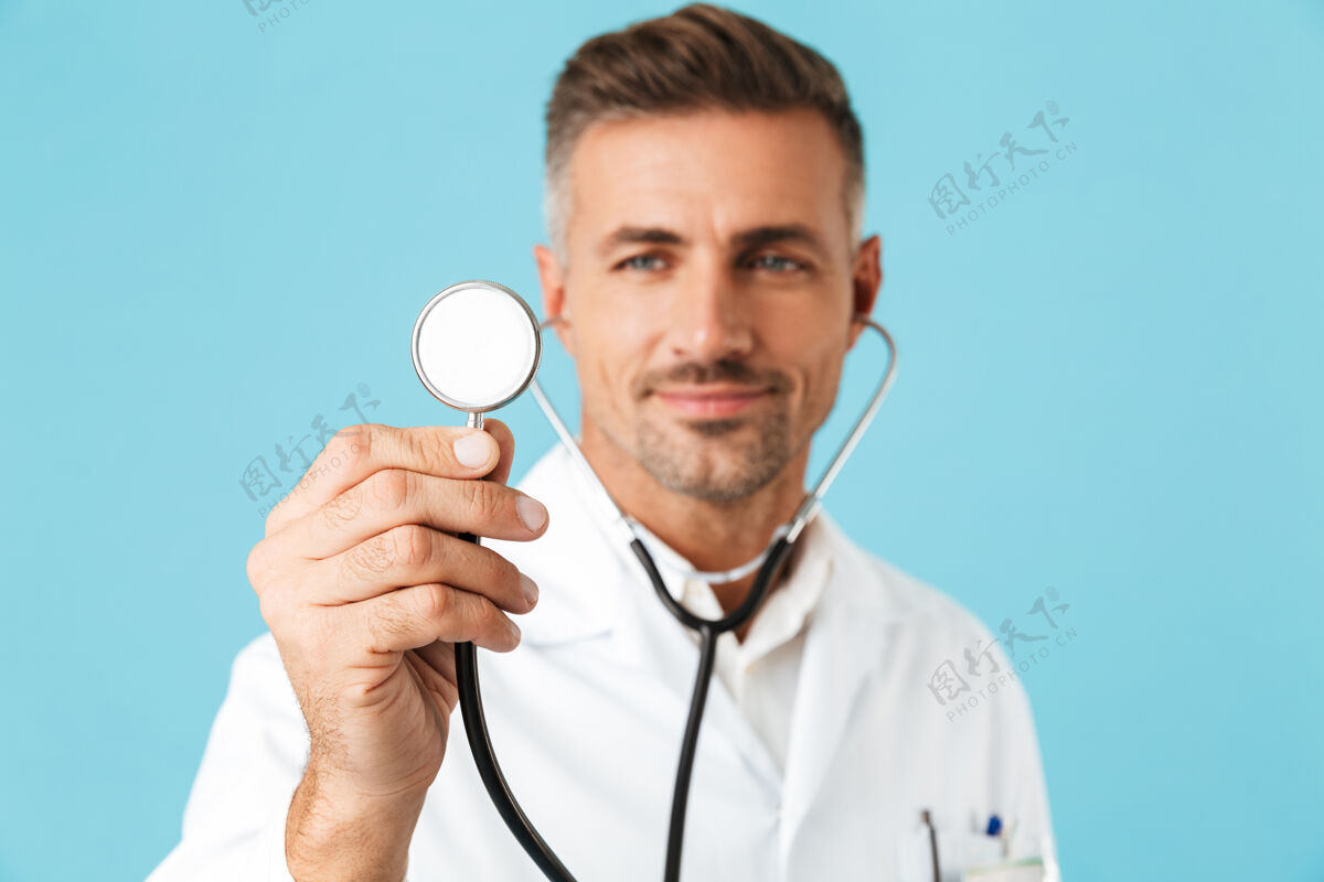 表情中年医生身穿白色制服手持听诊器 孤零零地站在蓝色墙壁上的画像医生成人男人