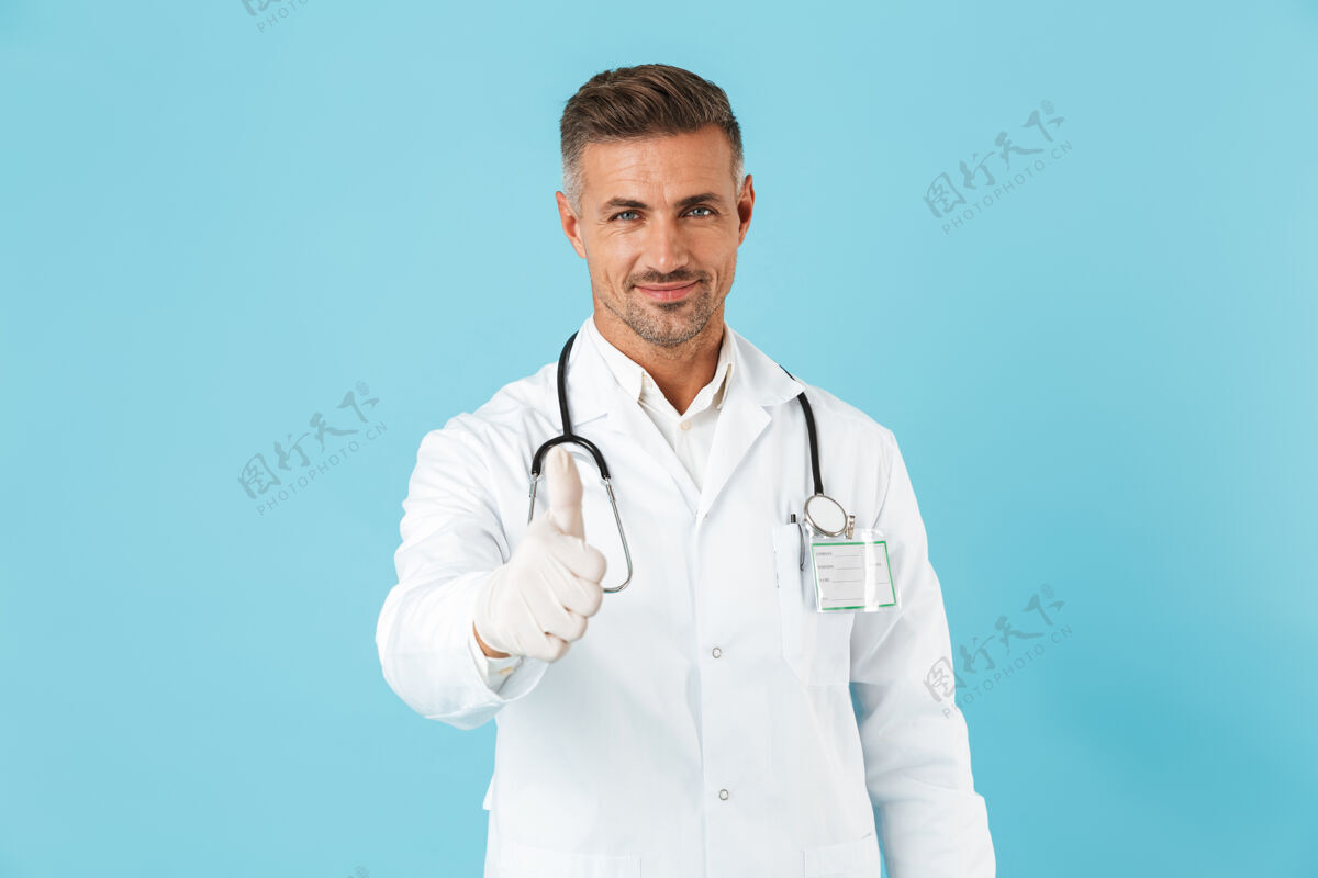 专家一个英俊的成熟男子医生在蓝色的墙上摆姿势 竖起大拇指东西中年工人