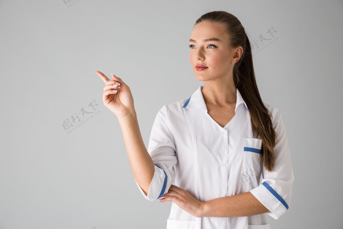 肖像一位年轻漂亮的美容师医生的照片被隔离在灰色的墙上指着制服女人医学