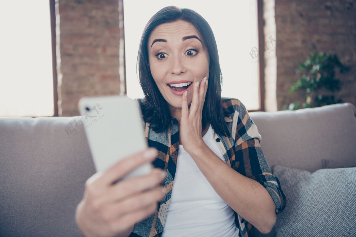 互联网美女手持电话阅读新instagram的照片发表正面评论喜出望外坐在舒适的沙发上穿着休闲服公寓室内衬衫手机休闲