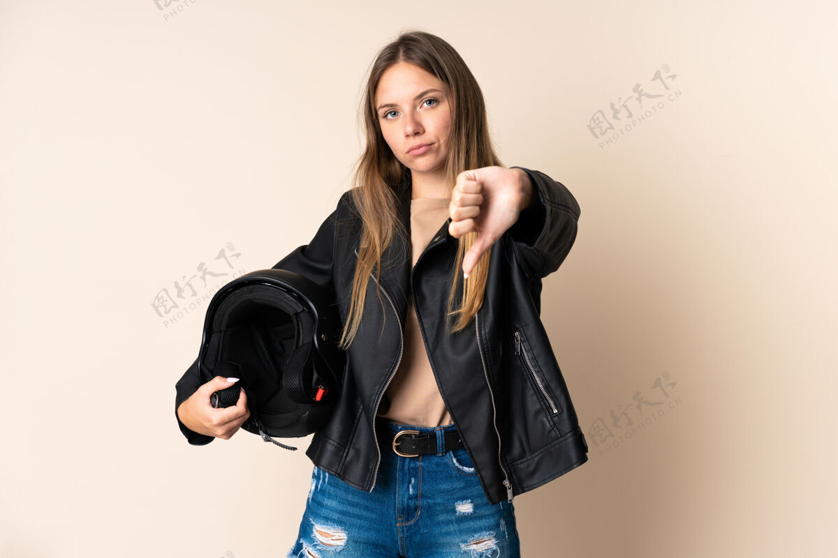 不同意立陶宛年轻女子手持摩托车头盔 背景为米色 拇指朝下 表情消极表情女孩支架