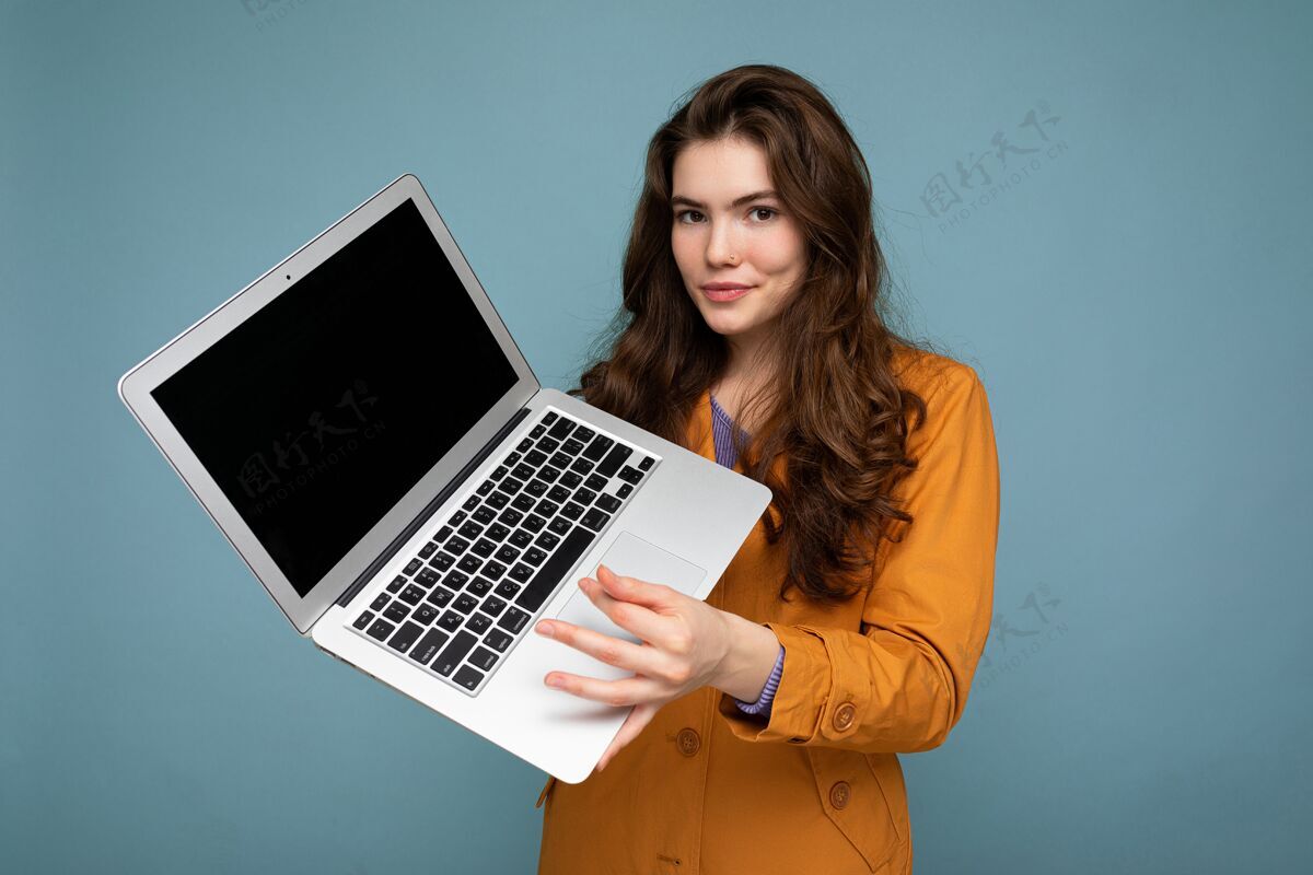技术照片中 美丽严肃快乐的年轻女子手持笔记本电脑 穿着黄色夹克 隔着蓝色的墙壁看着摄像机背景.断流器!技术 计算机 教育 学生 笔记本电脑 工作 数字 互联网 学习 游戏 工作 技术 信息 在线 电子 游戏 商务 无线 职业 便携式 上网本