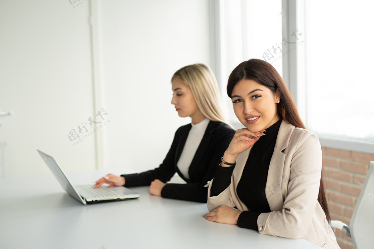 中心两个漂亮的年轻女子坐在办公室的桌子旁 手里拿着一台笔记本电脑笔记本会议商务