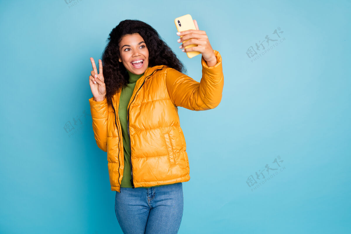 民族时髦的深色皮肤卷曲女士拿着电话自拍显示v符号穿时髦的黄色秋季外套牛仔裤绿色毛衣蓝色隔离墙微笑电话女性