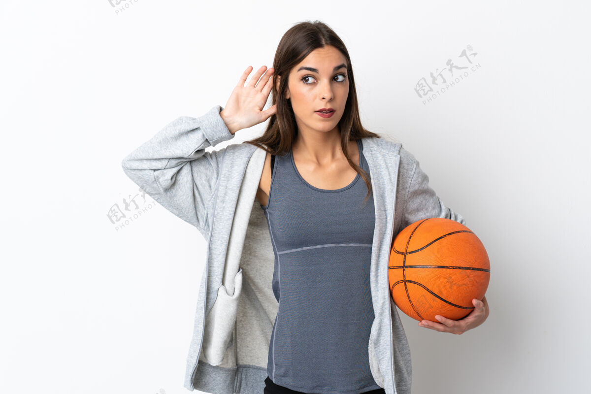 八卦在白色背景下孤立地打篮球的年轻女子把手放在耳朵上听东西女孩注意力年轻
