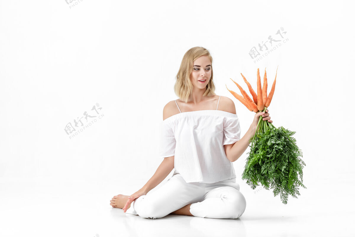 束一个穿着白衬衫的金发美女 手里拿着一根新鲜的胡萝卜 上面有绿叶背景.健康还有节食吃脸饮食