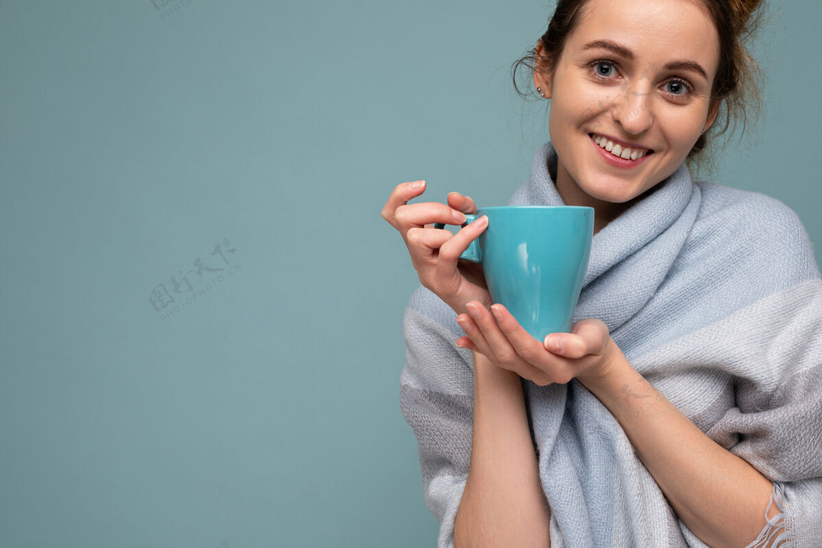 咖啡这张特写照片拍摄的是一位年轻漂亮 快乐 微笑的黑发女子 她戴着温暖的蓝色围巾 隔离在蓝色背景下 手持蓝色马克杯 喝着咖啡 看着我照相机.副本空间甜的可口的奶昔