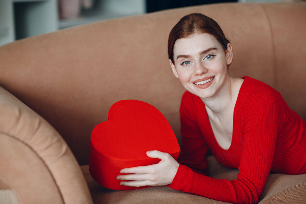 年轻美丽的红发女青年躺在客厅的沙发上 手里拿着心形礼品盒微笑情人节礼物今天还是生日浪漫白色床