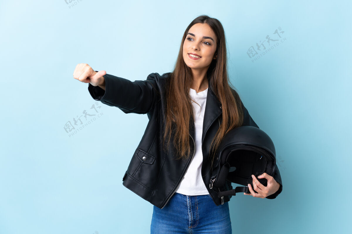 积极年轻的高加索女子手持摩托车头盔孤立在蓝色背景下竖起大拇指的手势手势好表情