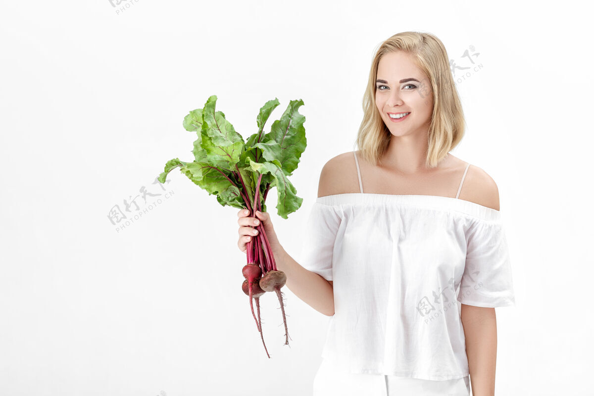 年轻一位穿着白色上衣的金发美女手持一根长着绿叶的甜菜根背景.健康还有维生素叶子饮食挑选