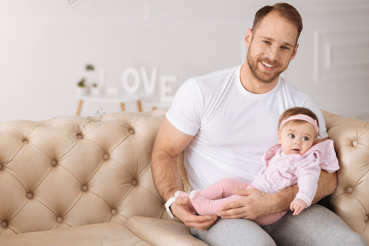 团结快乐的年轻人父亲体格健壮笑容可掬的大胡子男人坐在家里的沙发上 抱着自己的宝宝 同时表达着积极的心情童年服务沙发