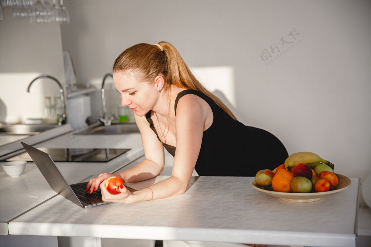 胃年轻的金发孕妇穿着黑色连衣裙在白色厨房里吃水果 看着我笔记本电脑高高质量的照片笔记本电脑腹部咨询