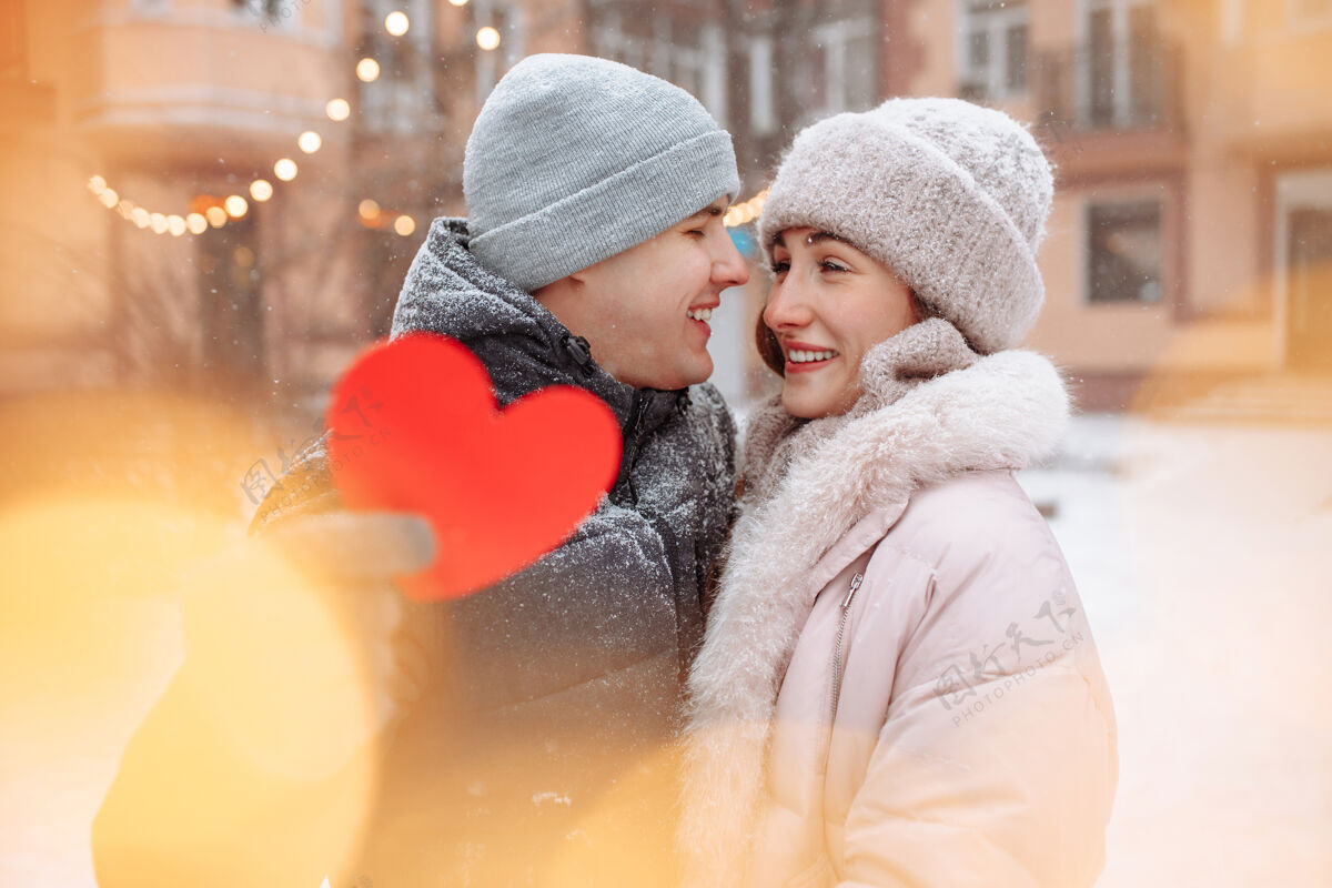 女朋友情人节的概念 相爱的情侣在冬天的雪地里亲吻拥抱帕克·杨男人拿着一个红色的纸心和他的女朋友一起庆祝情人节一对情侣在一起感觉很温暖心举行天