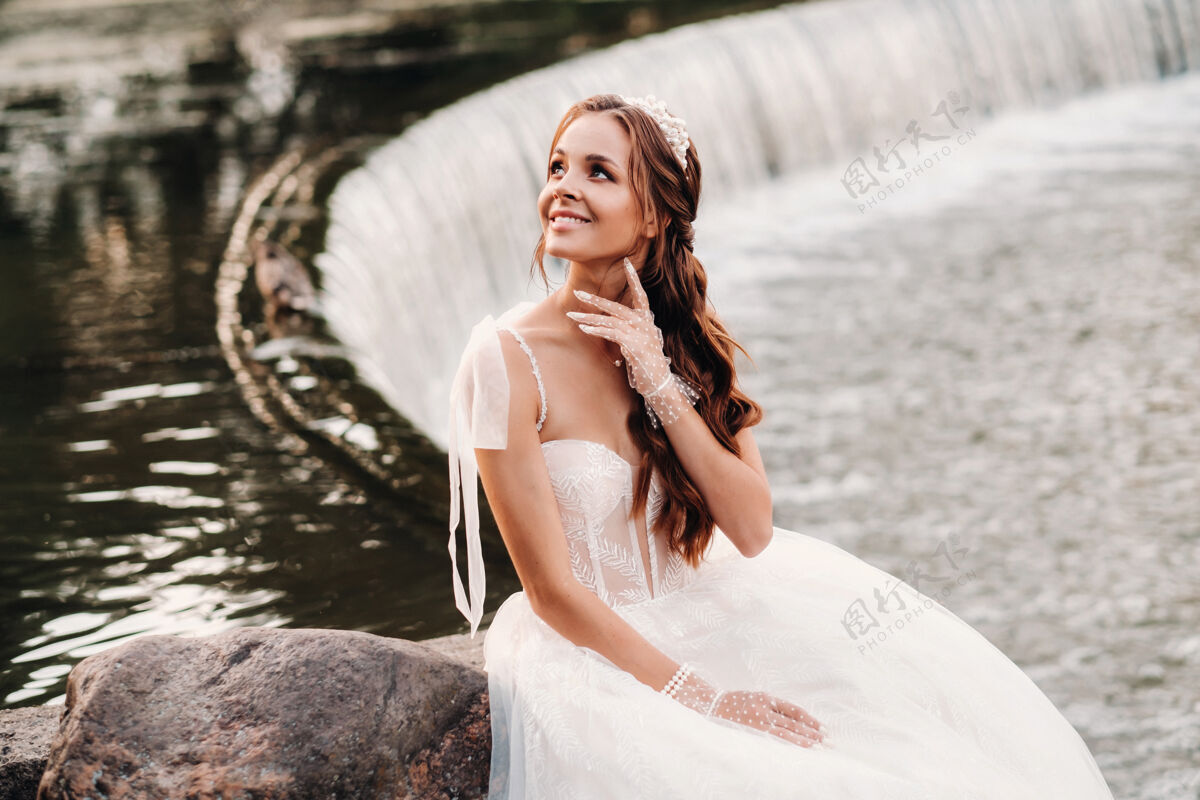 金发一位穿着白色礼服 戴着手套 赤脚的优雅新娘正坐在公园的瀑布旁 享受着大自然坐着微笑男人