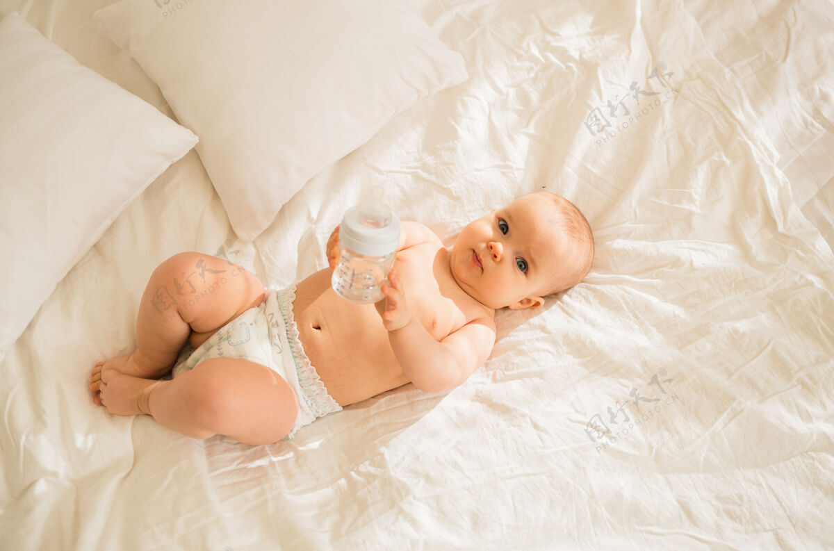坐着一个女婴躺在尿布里 床上的白色床单上放着一瓶水床婴儿饮料