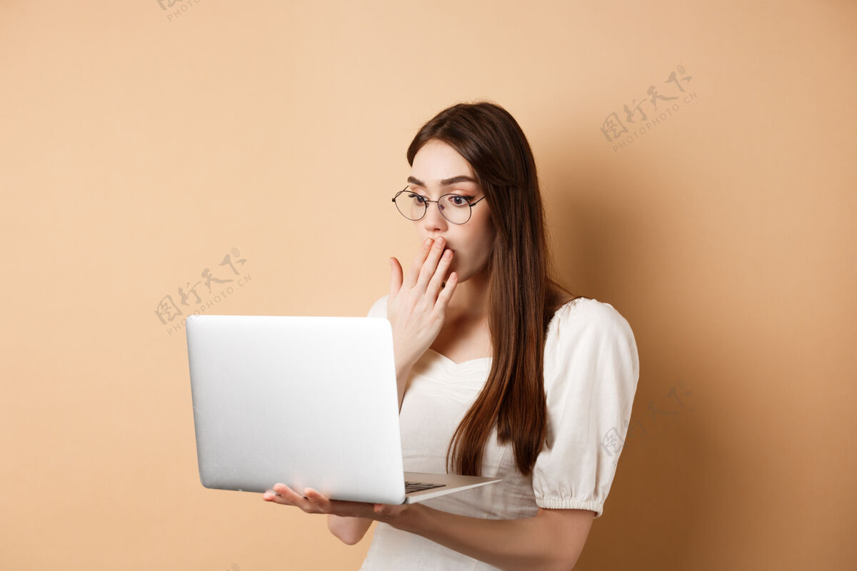 时尚震惊的女孩看着笔记本电脑屏幕 用惊讶的脸捂住嘴巴 站在米色眼镜上购物科技欢呼
