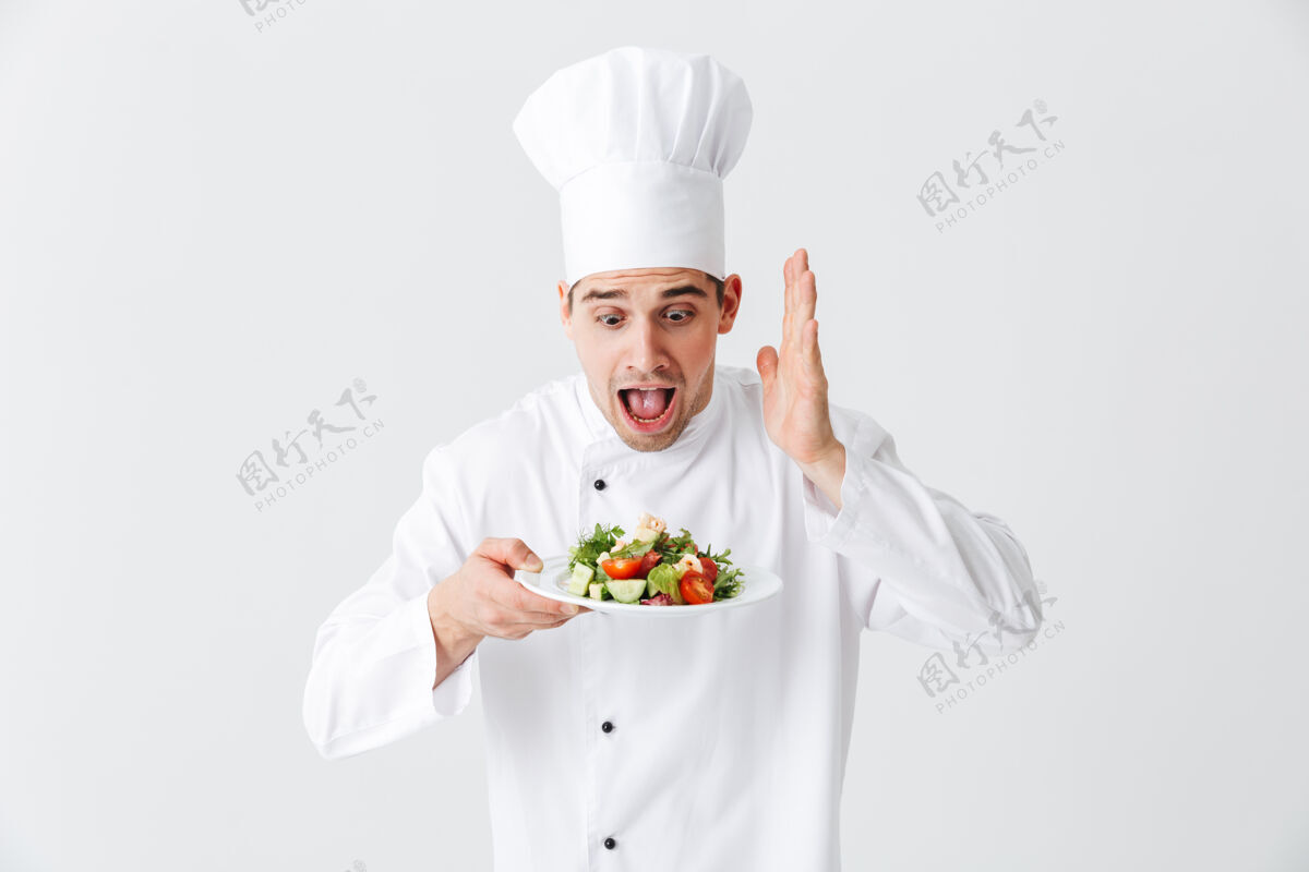 烹饪兴奋的男厨师穿着制服 把新鲜的蔬菜沙拉放在隔离在白色墙上的盘子上举行制服膳食