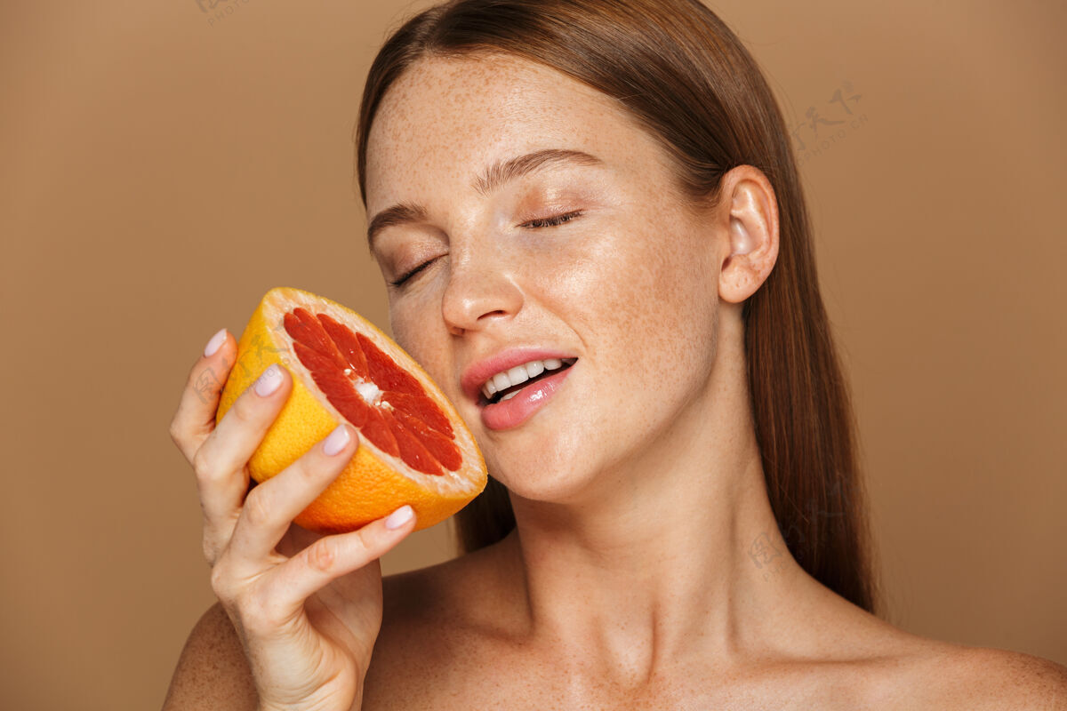 葡萄柚美丽的肖像 一个美丽健康的年轻裸体女子长着红色的头发显示切片柚子隔离在米色墙上年轻纯洁白种人